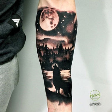 Black and white forearm tattoo of a wolf on the bank of a moon-light river howling towards the moon with fir trees and a hill