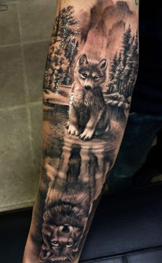 Forearm black and white tattoo of a wolf cub looking down at a reflection of its older self in stream with a tree background